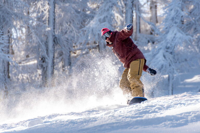Snowboarder con giacca bordeaux in curva nella neve fresca tra gli alberi innevati di Sestriere, servizio fotografico sportivo sulla neve