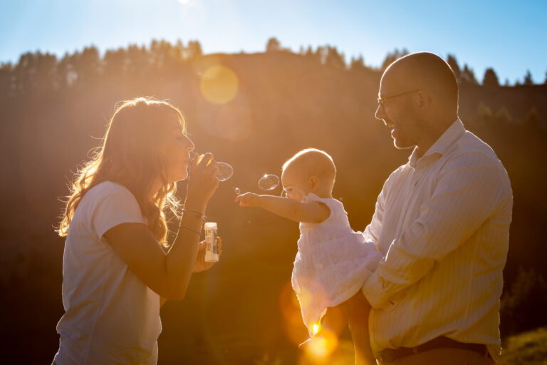 Mamma e papà con la bambina in braccio che gioca con le bolle di sapone, in controluce al tramonto in montagna, servizio fotografico di famiglia in Alta Val di Susa